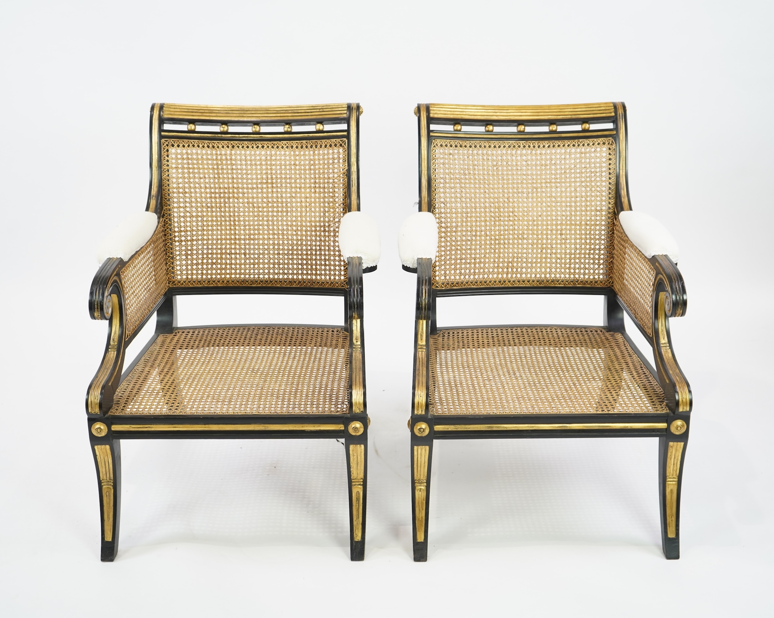 A pair of Regency style ebonised, parcel gilt caned bergere elbow chairs